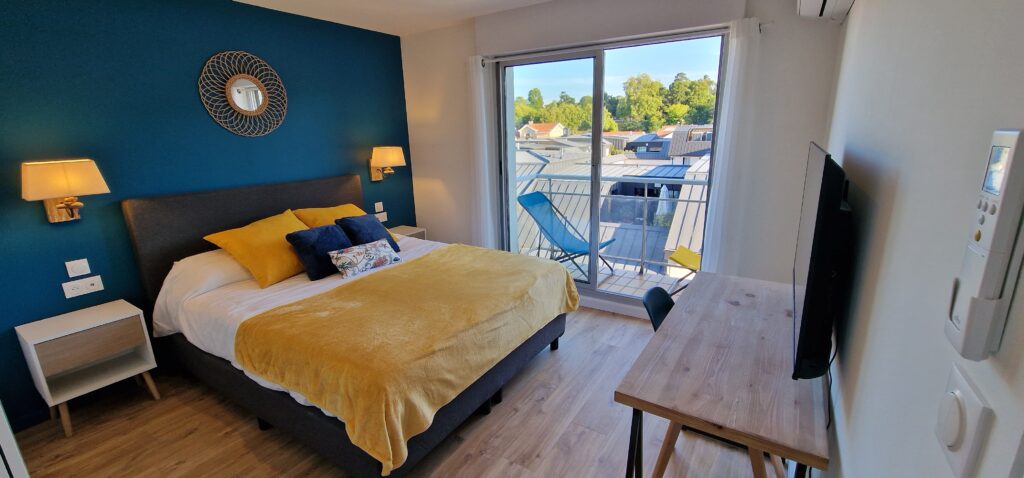 Une chambre blanche avec un lit 160cm surplombant un mur de couleur bleue océan. Un accès balcon est visible, avec une vue sur les toits d'Arcachon. 