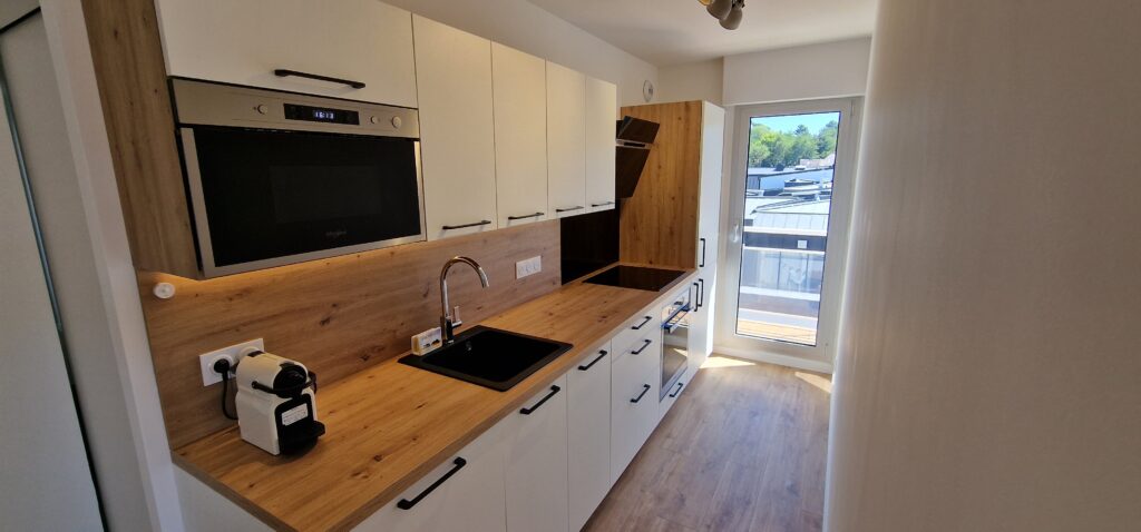 Une cuisine blanche avec des surfaces de style bois. Un accès balcon est visible. 