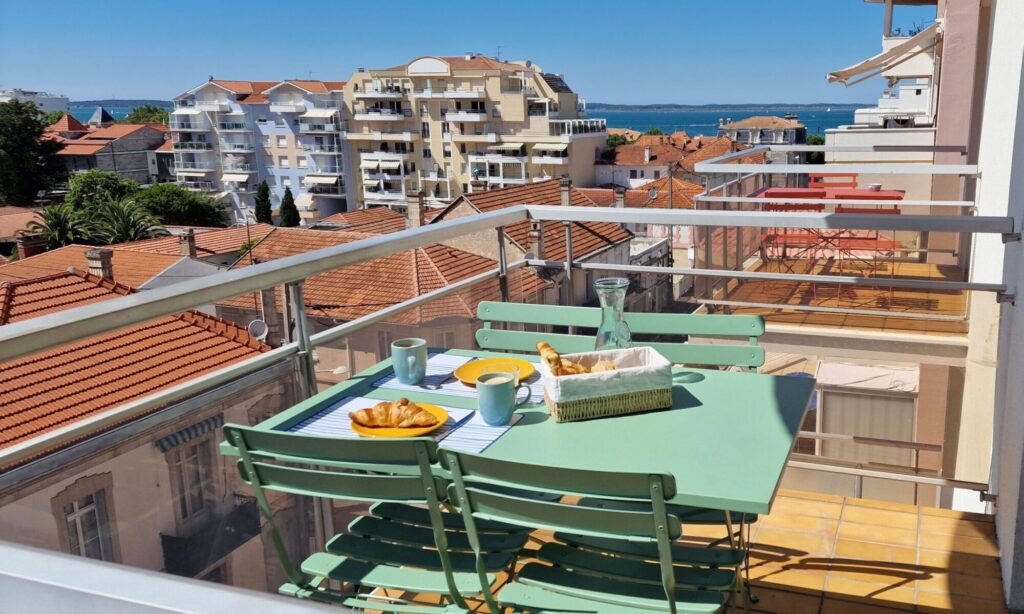 Une terrasse avec vue sur la mer. Un petit-déjeuner se trouve sur une table verte avec quatre chaises qui l'entourent. 